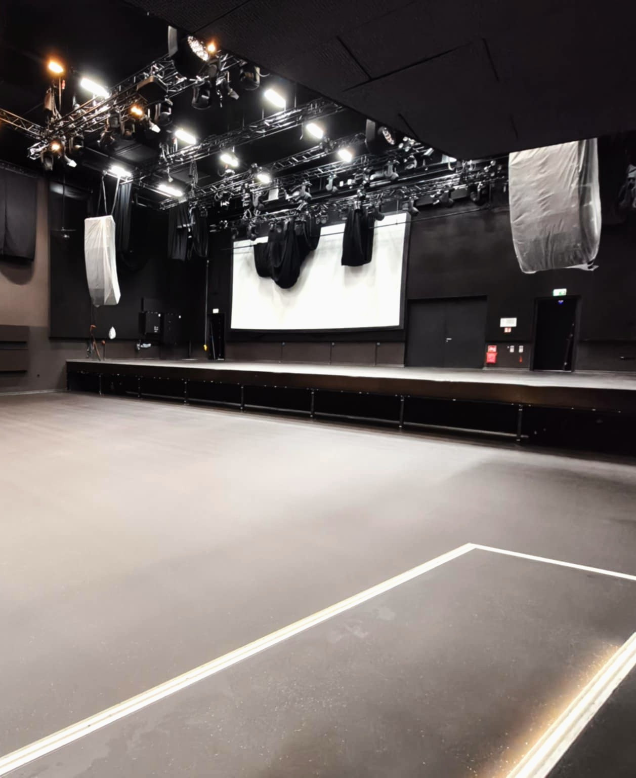Szara, gładka posadzka w dużej sali widowiskowej z oświetleniem technicznym. Widoczne białe linie na podłodze i czarne ściany. Scena z ekranem w tle.