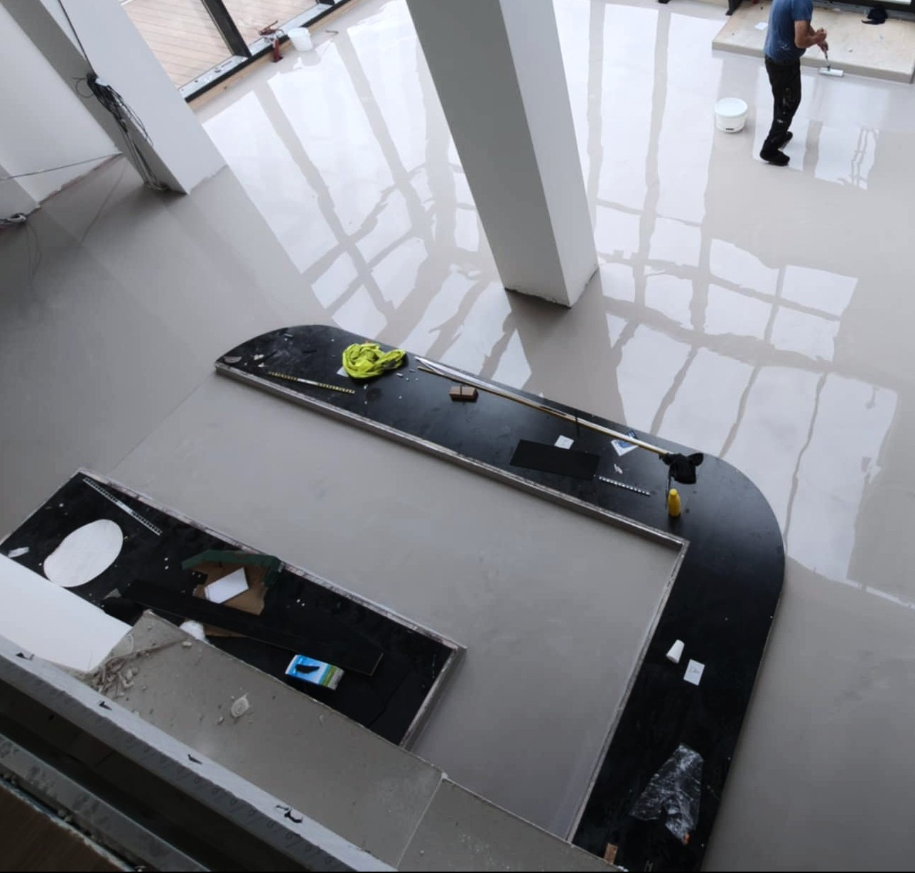 Wylewanie jasnej posadzki żywicznej w przestronnym wnętrzu. Widoczne narzędzia pracy i odbicia światła w mokrej powierzchni. Praca w toku, widok z góry.