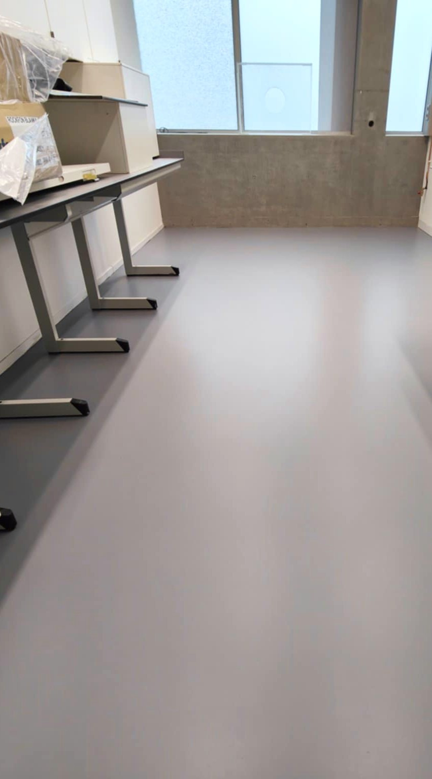 Gładka, szara posadzka w jasnym pomieszczeniu z oknami i betonową ścianą. Widoczne fragmenty metalowych nóg stołów. Minimalistyczny, nowoczesny design.