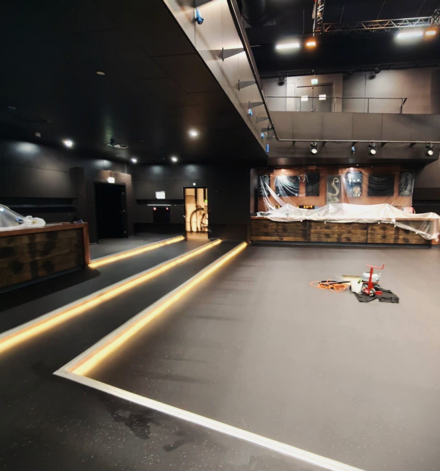 Ciemna, gładka posadzka z wbudowanym oświetleniem liniowym w lokalu usługowym. W tle bar zabezpieczony folią i narzędzia pracy.