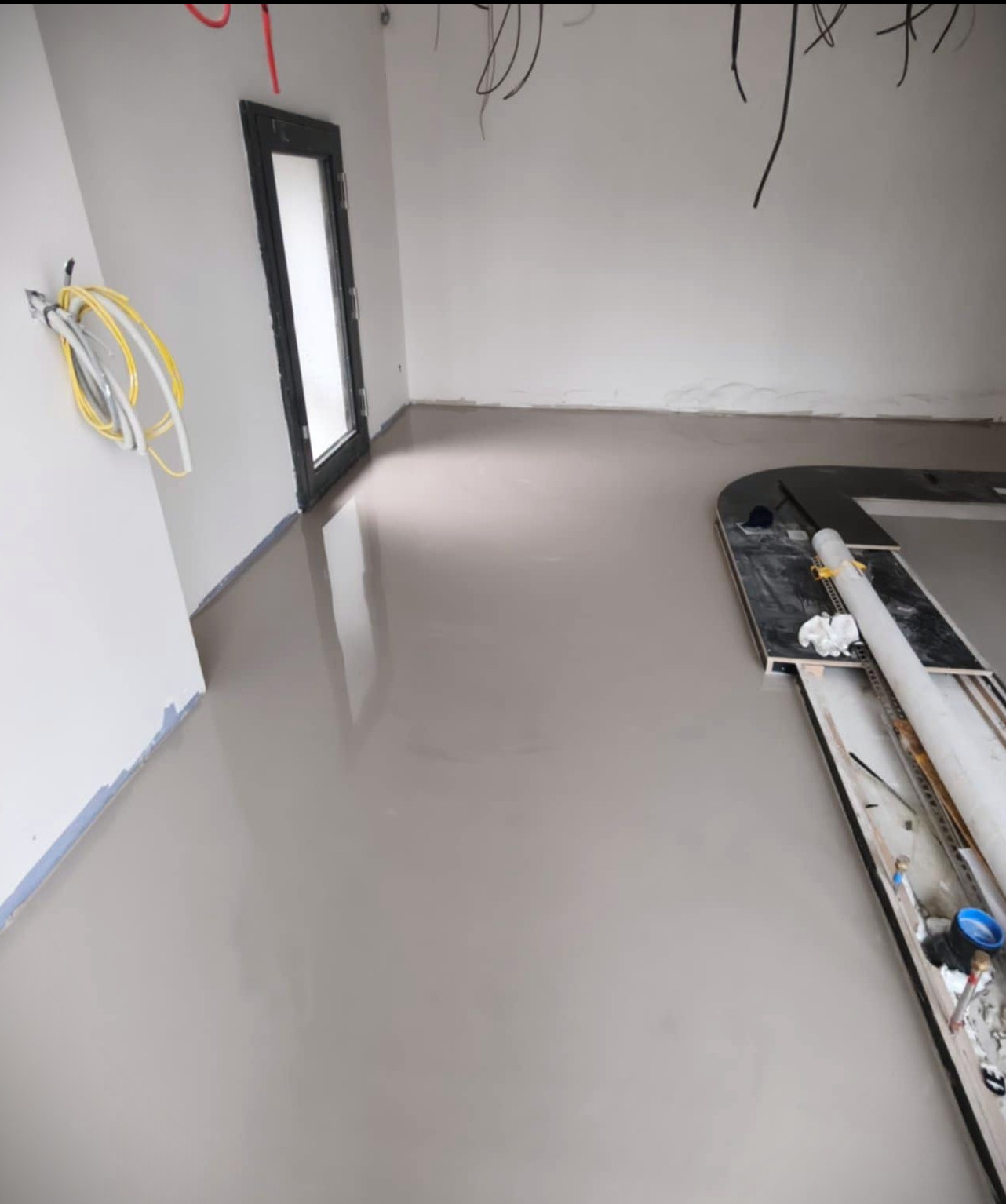Gładka, szara posadzka w surowym wnętrzu z widocznymi przewodami i narzędziami, świeżo po wylaniu, z czarną ramą drzwi w tle. Minimalistyczny, nowoczesny styl.