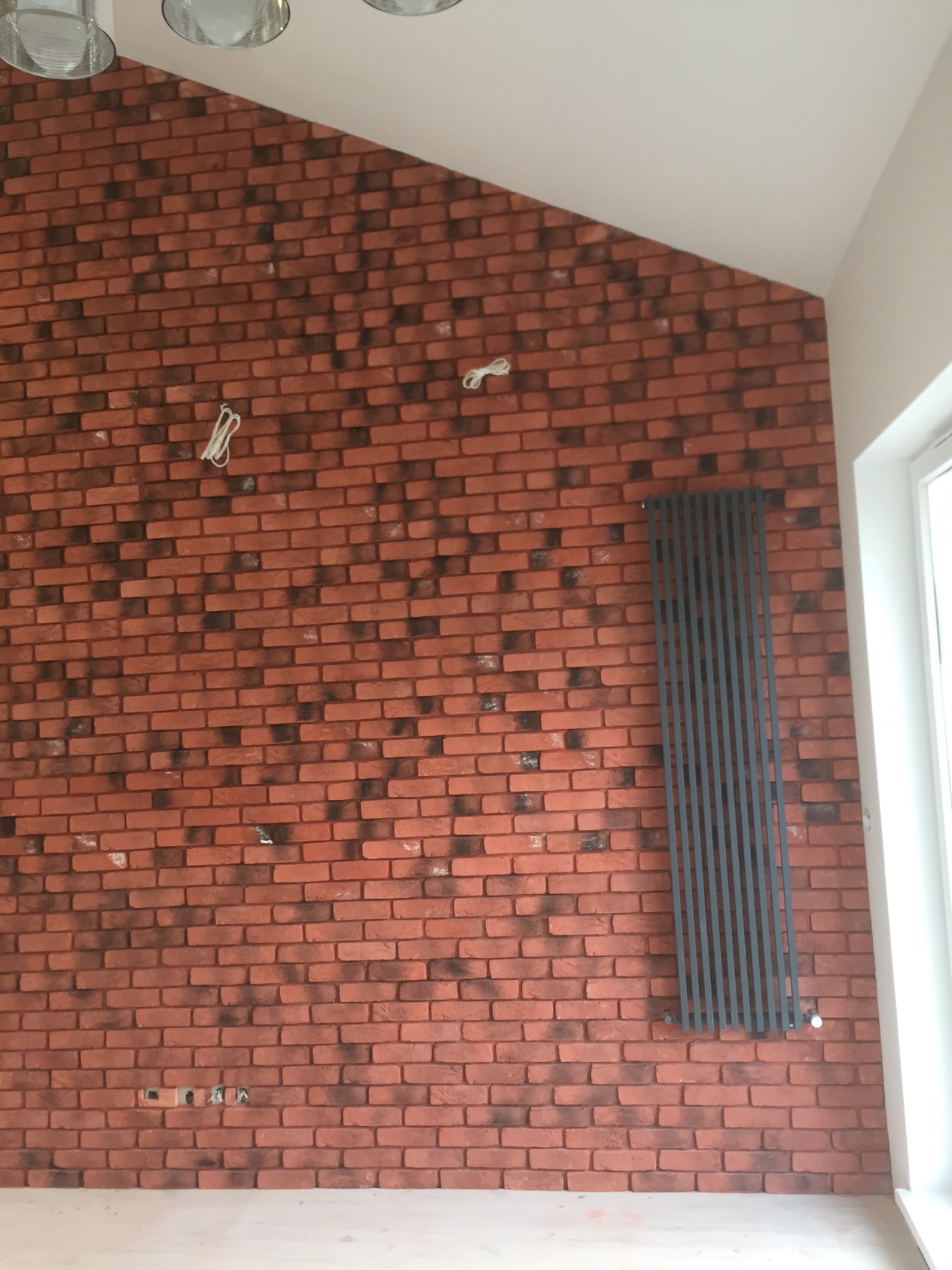 Ściana z cegły klinkierowej w odcieniach czerwieni i brązu, z zamontowanym szarym grzejnikiem panelowym i wystającymi przewodami elektrycznymi, oświetlona światłem dziennym w narożniku pomieszczenia.