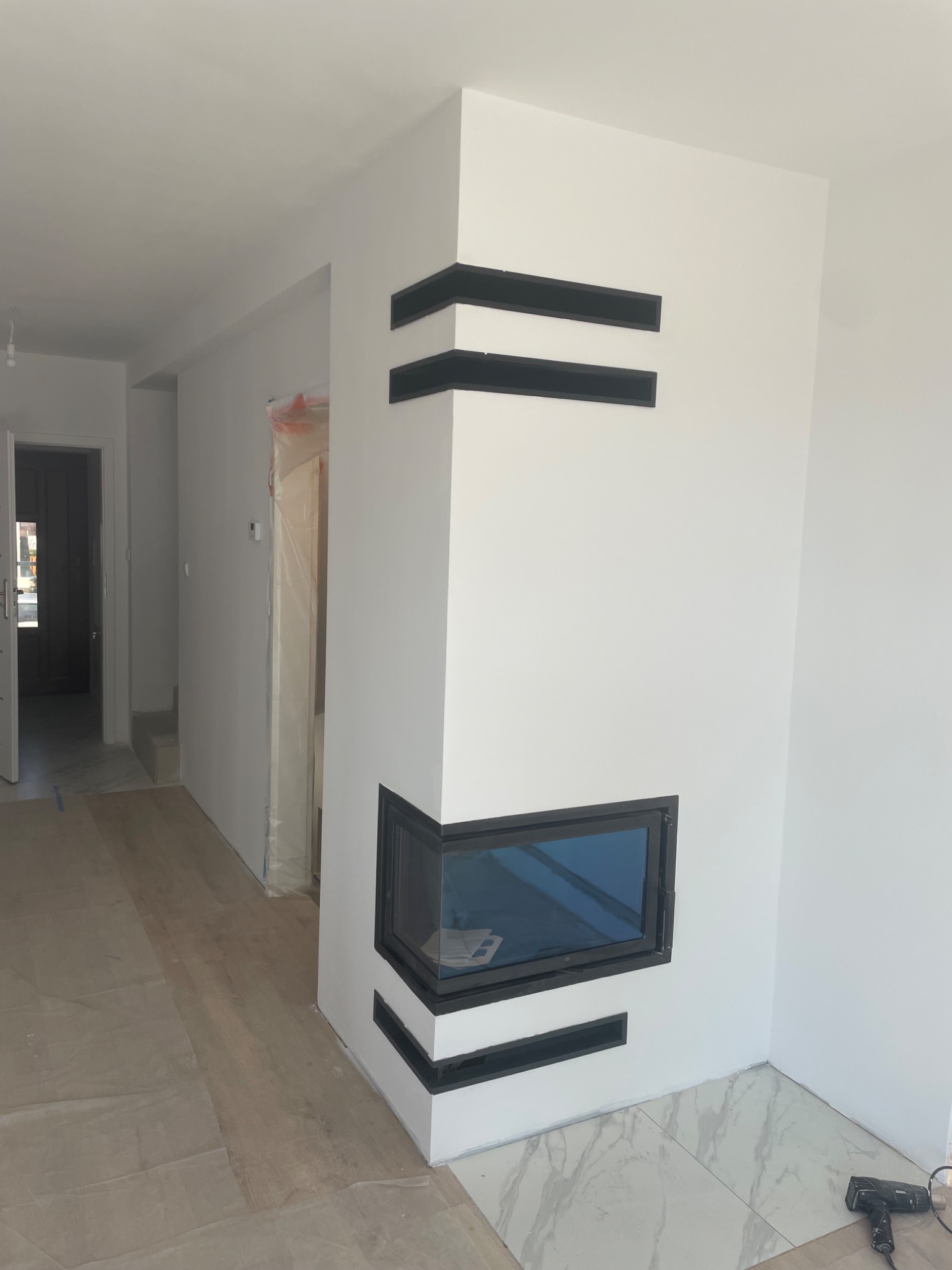 Nowoczesny kominek narożny z czarnymi, poziomymi zdobieniami na białej ścianie, widok z boku, podłoga częściowo wyłożona marmurowymi płytkami, częściowo zabezpieczona folią ochronną, w tle widoczne...