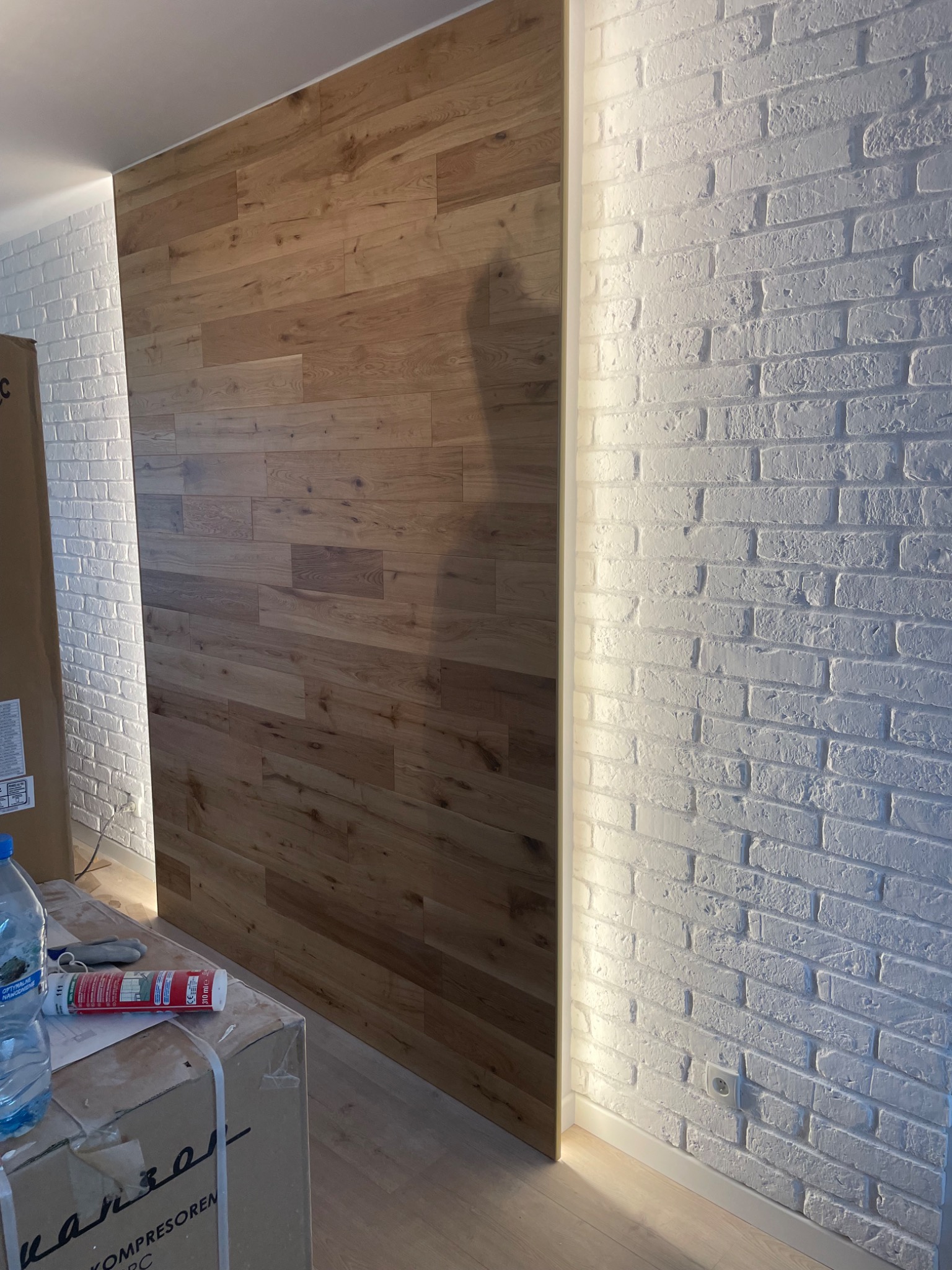 Wykończona ściana z paneli drewnianych sąsiadująca z białą ścianą z cegły dekoracyjnej, oświetlona listwą LED, widoczny fragment kartonu z akcesoriami montażowymi.