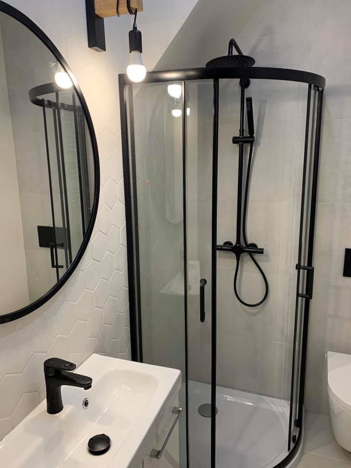 Nowoczesna łazienka z czarnymi akcentami: kabina prysznicowa narożna, okrągłe lustro z czarną ramą, biała umywalka z czarną baterią i heksagonalne płytki na ścianie.