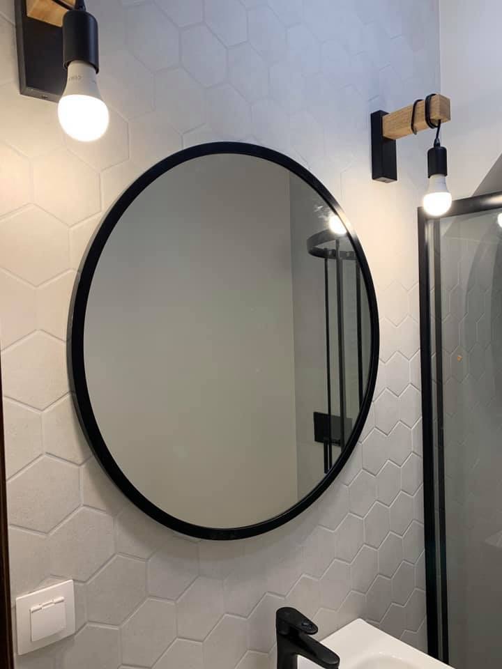 Okrągłe lustro w czarnej ramie na ścianie wyłożonej białymi heksagonalnymi płytkami; widoczne minimalistyczne oświetlenie z drewnianymi elementami i czarnymi oprawkami, bateria umywalkowa w kolorze...