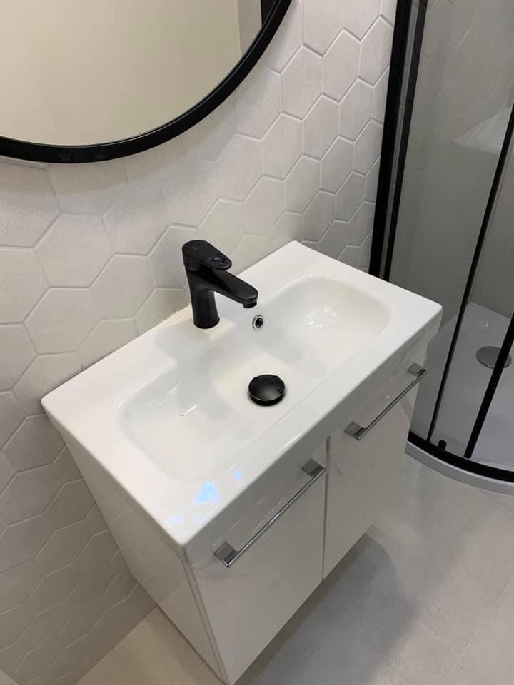 Nowoczesna łazienka z białą szafką pod umywalkę, czarną baterią i okrągłym lustrem na ścianie wyłożonej heksagonalnymi płytkami, kabina prysznicowa z czarnymi ramami w tle.