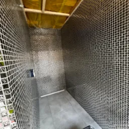 Wnętrze łazienki w trakcie remontu z błyszczącą mozaiką na ścianach, szarymi płytkami na podłodze i widoczną konstrukcją sufitu oraz narzędziami malarskimi w rogu.