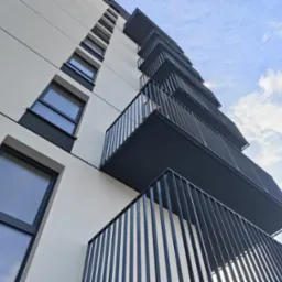 Nowoczesny budynek z balkonami i metalowymi balustradami, widok z dołu na tle błękitnego nieba z chmurami. Minimalistyczna architektura.