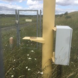 WELD-PROJECT - Żółta metalowa kolumna z przymocowaną skrzynką i półką, otoczona trawą i kwiatami, z metalową bramą w tle.