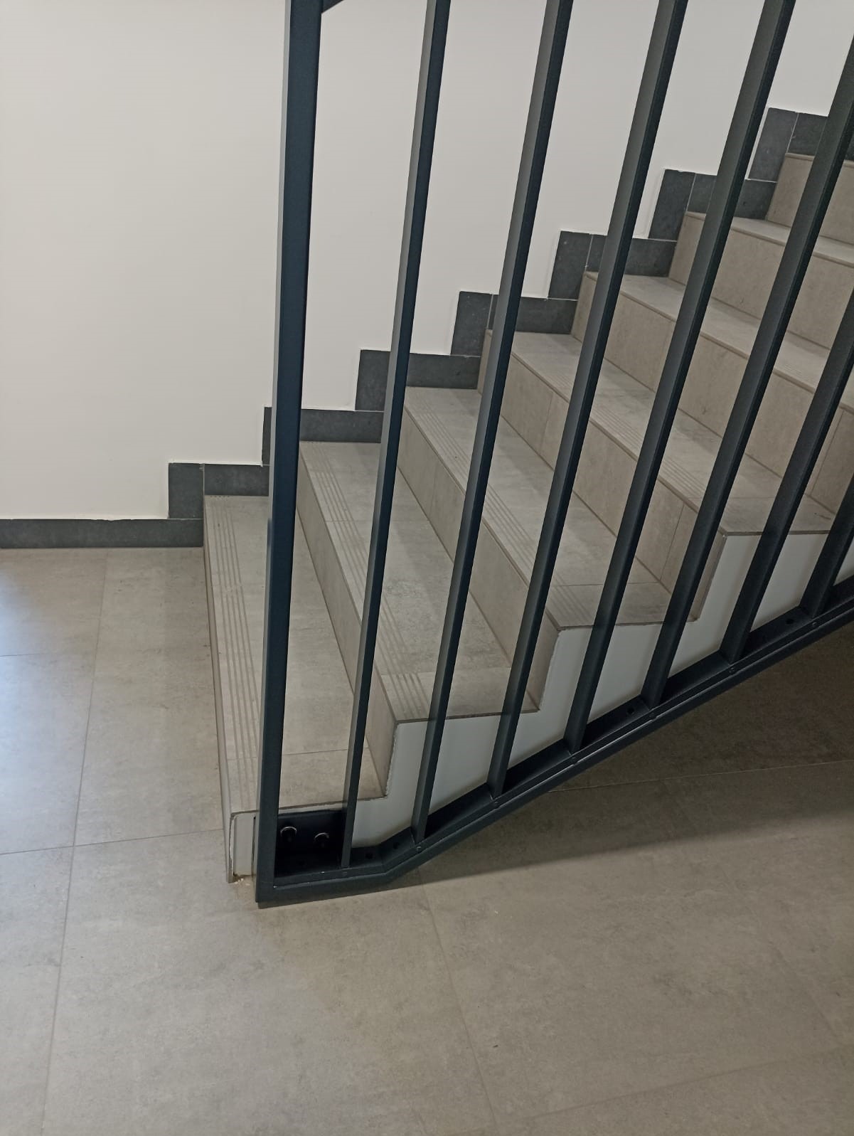 Wnętrze: schody z szarymi stopniami i metalową balustradą z pionowymi szczeblami. Widok z dołu, minimalistyczny design, neutralna kolorystyka.
