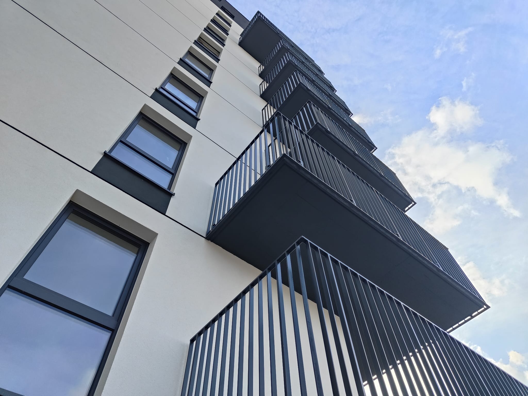 Nowoczesny budynek z balkonami i metalowymi balustradami, widok z dołu na tle błękitnego nieba z chmurami. Minimalistyczna architektura.
