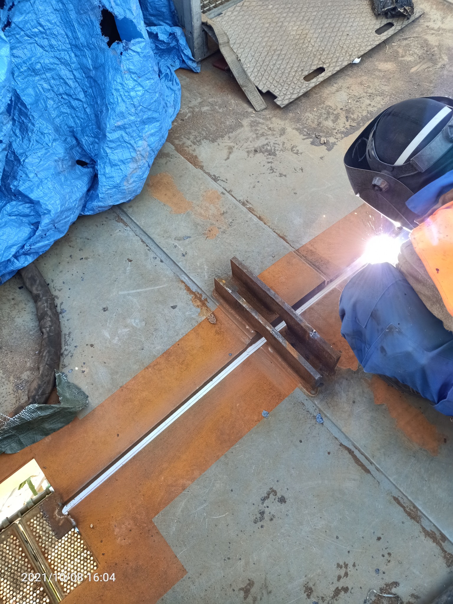 Spawanie metalowej konstrukcji z widocznym spawaczem w masce i iskrzącym spawem, na tle niebieskiej plandeki i metalowych elementów.