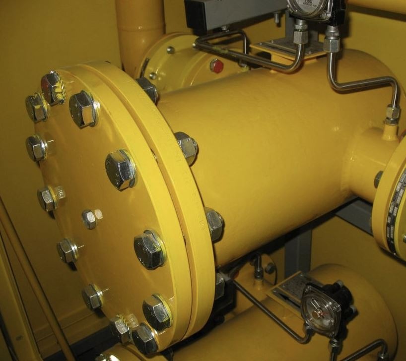 Żółta rura przemysłowa z licznymi śrubami mocującymi kołnierz, widoczne manometry i rurki. Specyficzna kolorystyka i detale przemysłowe.