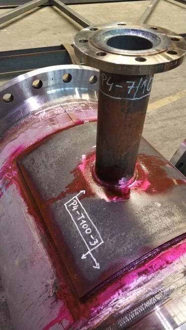 Spawanie rury do zbiornika z testem penetracyjnym w kolorze różowym. Widoczne białe oznaczenia na metalu. Fragment konstrukcji stalowej w hali.