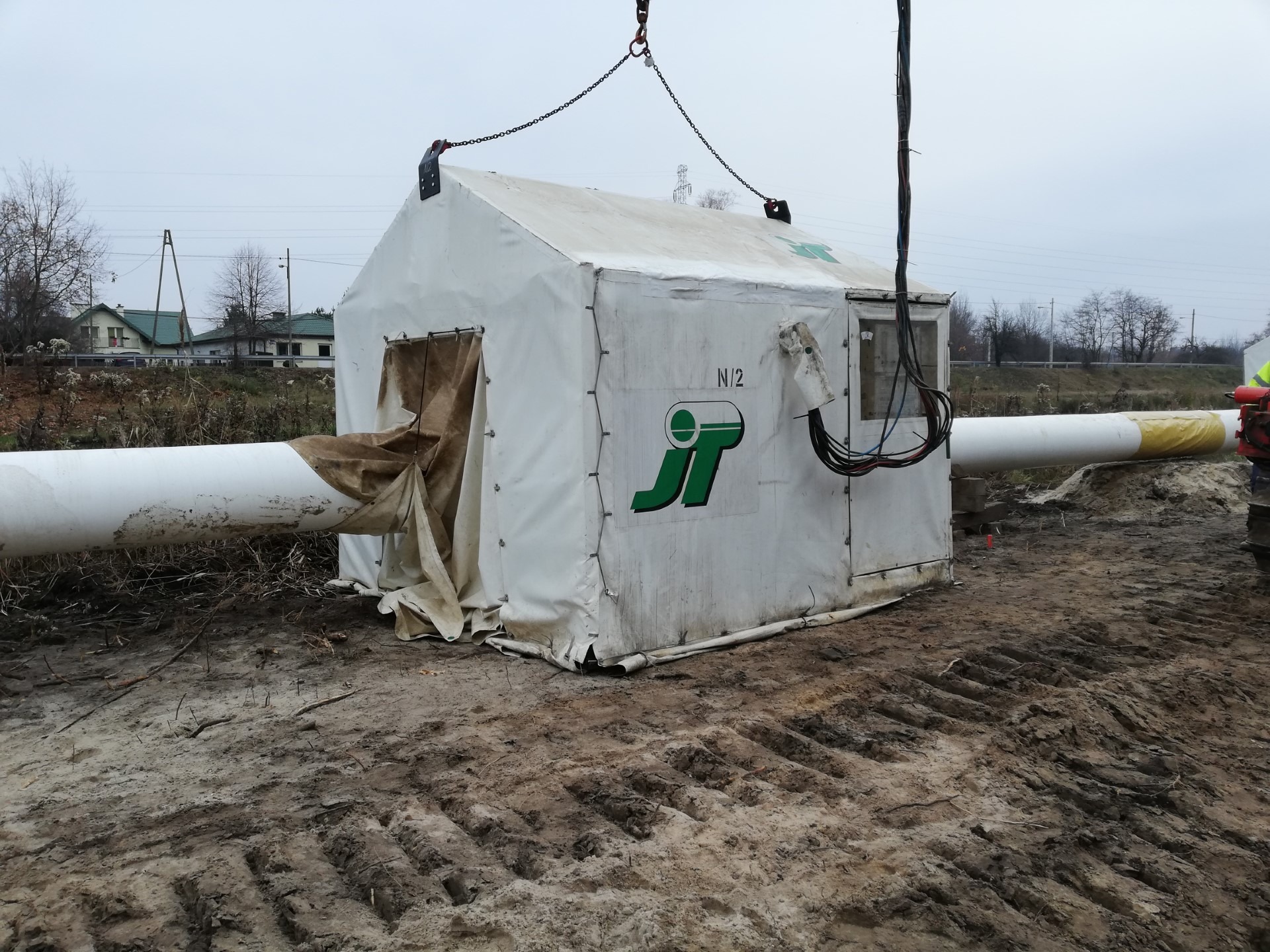 Mobilna komora spawalnicza z logo JT, zawieszona nad rurociągiem na placu budowy. Widoczne przewody i elementy konstrukcyjne, tło stanowią budynki i pochmurne niebo.