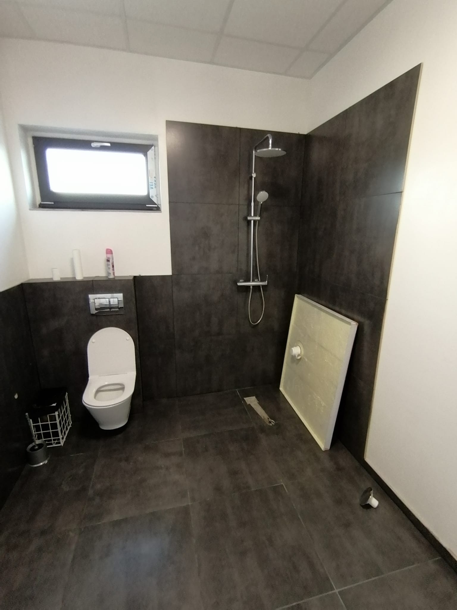 Nowa łazienka z ciemnymi płytkami, białą toaletą, kabiną prysznicową w trakcie montażu i oknem.