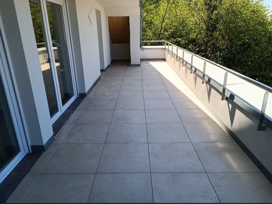 Jasnoszary taras wyłożony dużymi, kwadratowymi płytkami, z metalową balustradą i widokiem na zieleń, oświetlony popołudniowym słońcem tworzącym długie cienie.