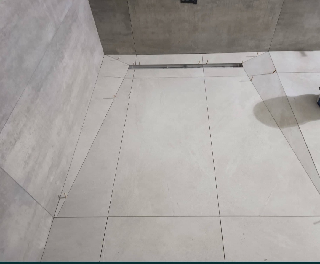 Ukończona podłoga prysznicowa z jasnoszarych, kwadratowych płytek, z widocznym odpływem liniowym i sznurkami wyznaczającymi spadek, po prawej widoczny fragment buta.