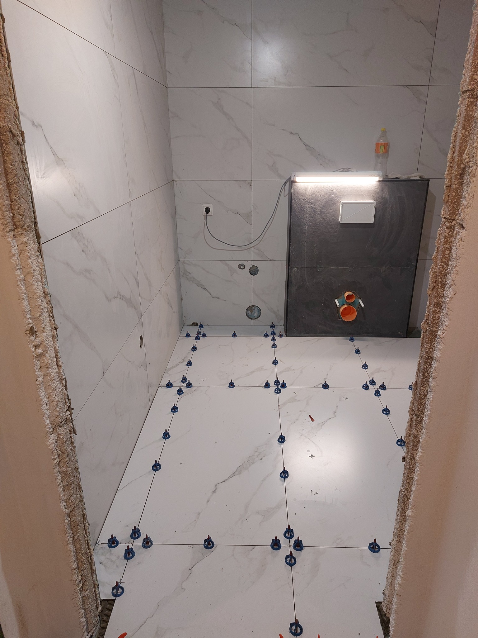 Montaż płytek imitujących marmur w łazience, widoczne niebieskie klipsy poziomujące, surowe ściany w tle, instalacja podtynkowego WC z oświetleniem i butelką wody na górze.