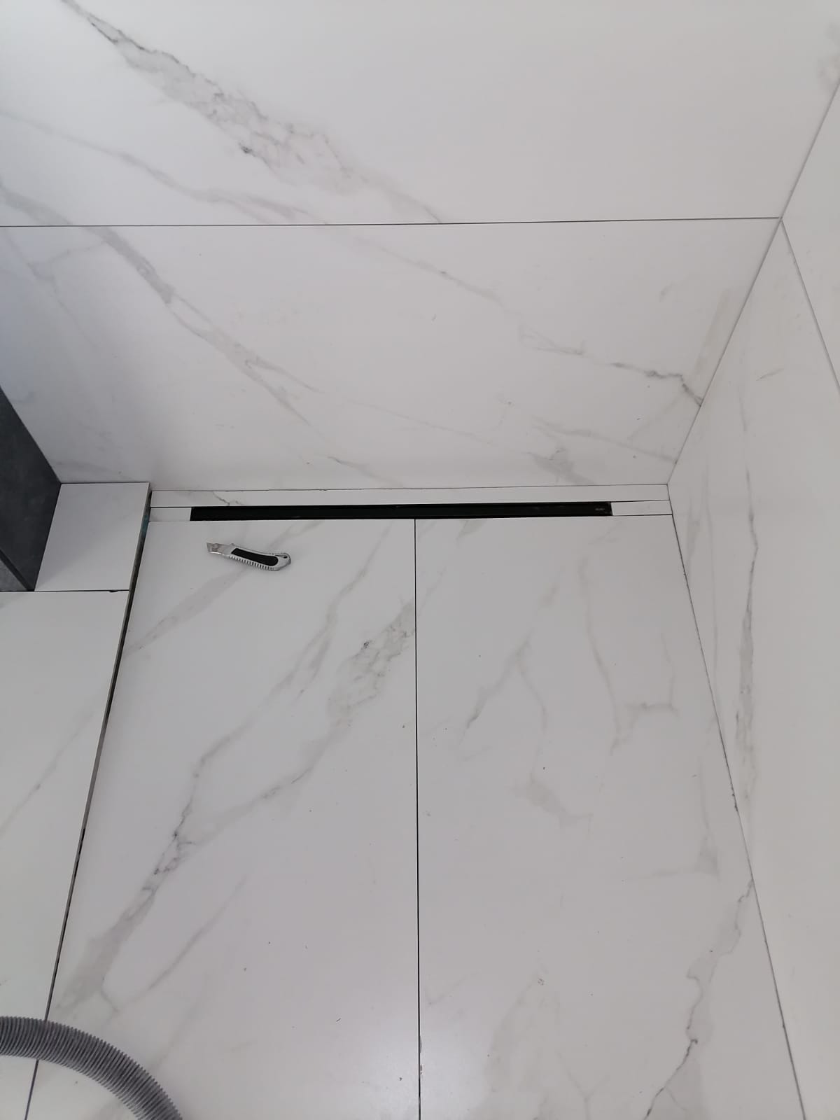 Białe płytki podłogowe z marmurkowym wzorem, czarny odpływ liniowy i nóż do cięcia płytek leżący na podłodze.