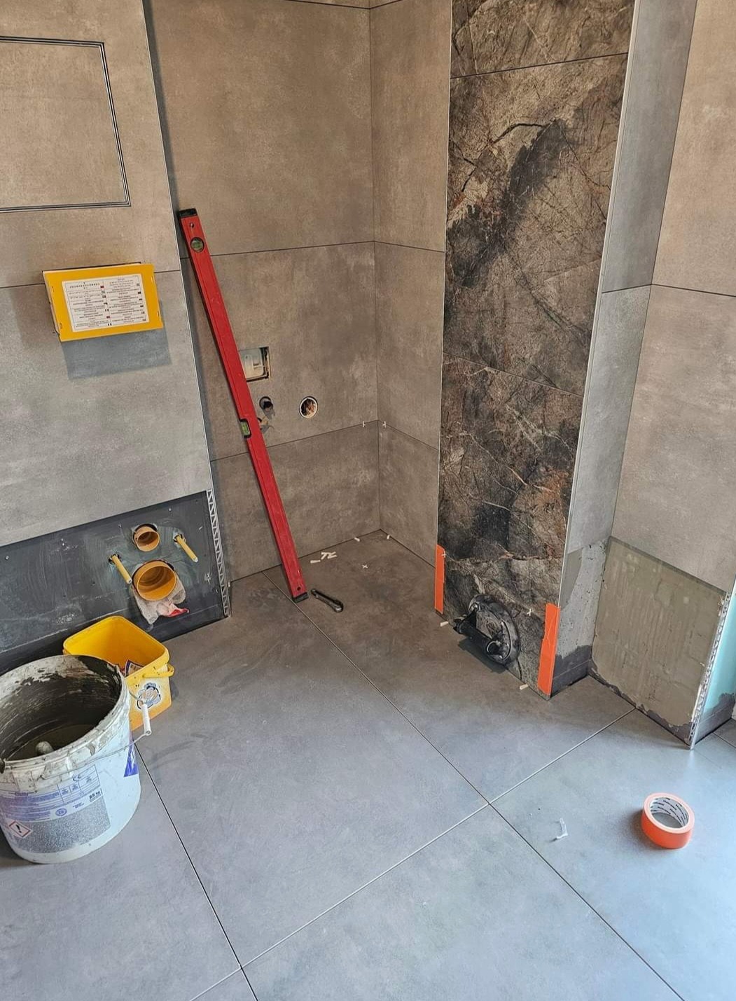 Montaż płytek w łazience: szare płytki podłogowe i ścienne, dekoracyjna płyta imitująca marmur, poziomica oparta o ścianę, wiadro z zaprawą, przygotowane przyłącza hydrauliczne.