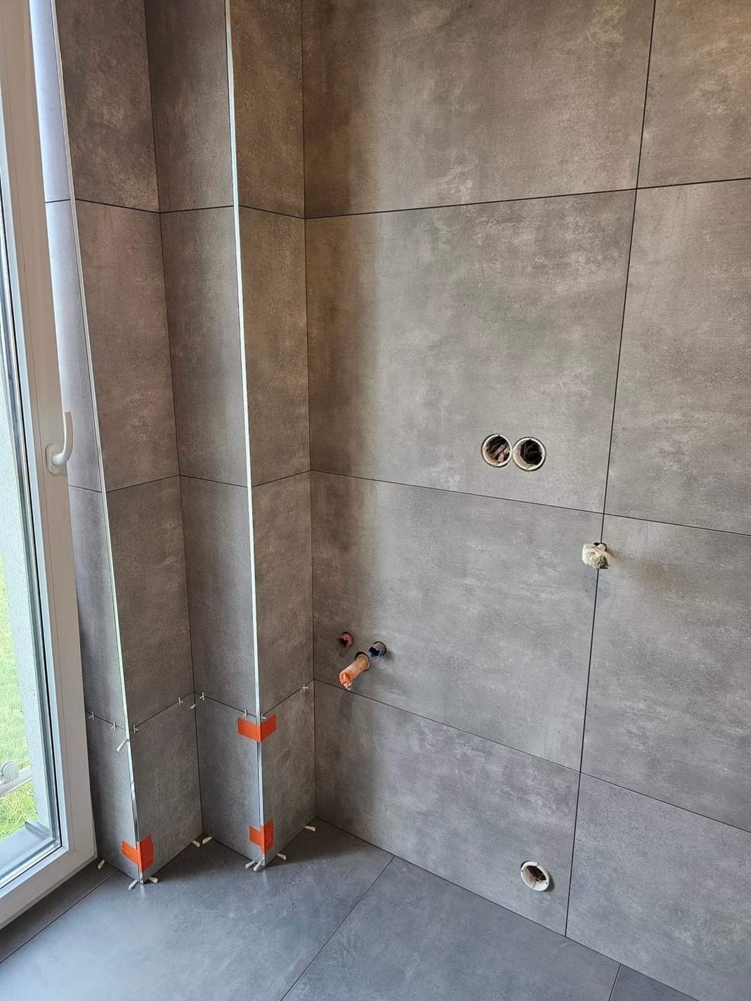 Szare płytki wielkoformatowe na ścianie łazienki w trakcie montażu, widoczne krzyżyki dystansowe i pomarańczowe narożniki zabezpieczające, przygotowane otwory na instalacje.