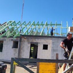 Małgorzata Moroz - Budowa domu: robotnicy montują drewnianą konstrukcję dachu na budynku z białego bloczka, widoczny dźwig i płyta OSB na ścianie szczytowej.