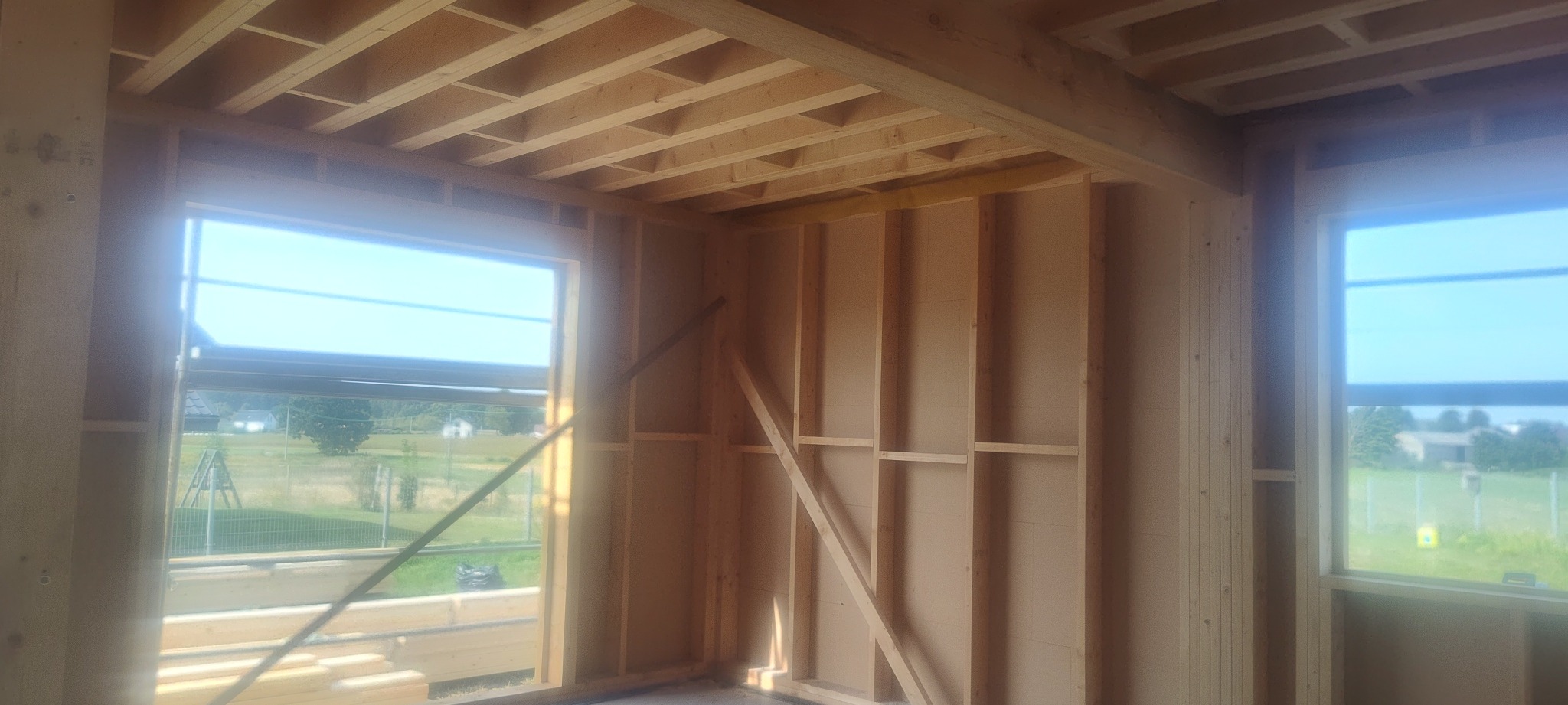 Wnętrze domu w trakcie budowy, widoczne drewniane ramy ścian, strop z belek, okna z widokiem na zewnątrz, w tle domy jednorodzinne.