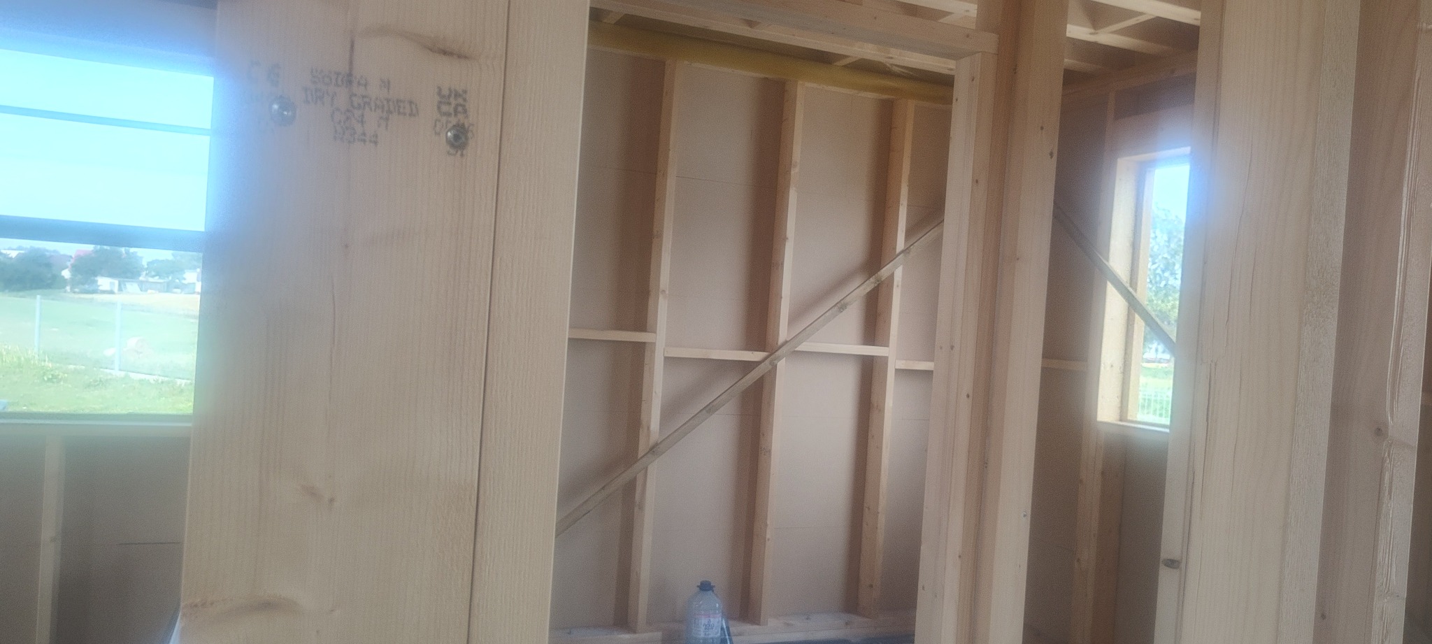 Wnętrze domu w trakcie budowy, widoczna drewniana konstrukcja szkieletowa ścian z oknami, butelka wody na podłodze.