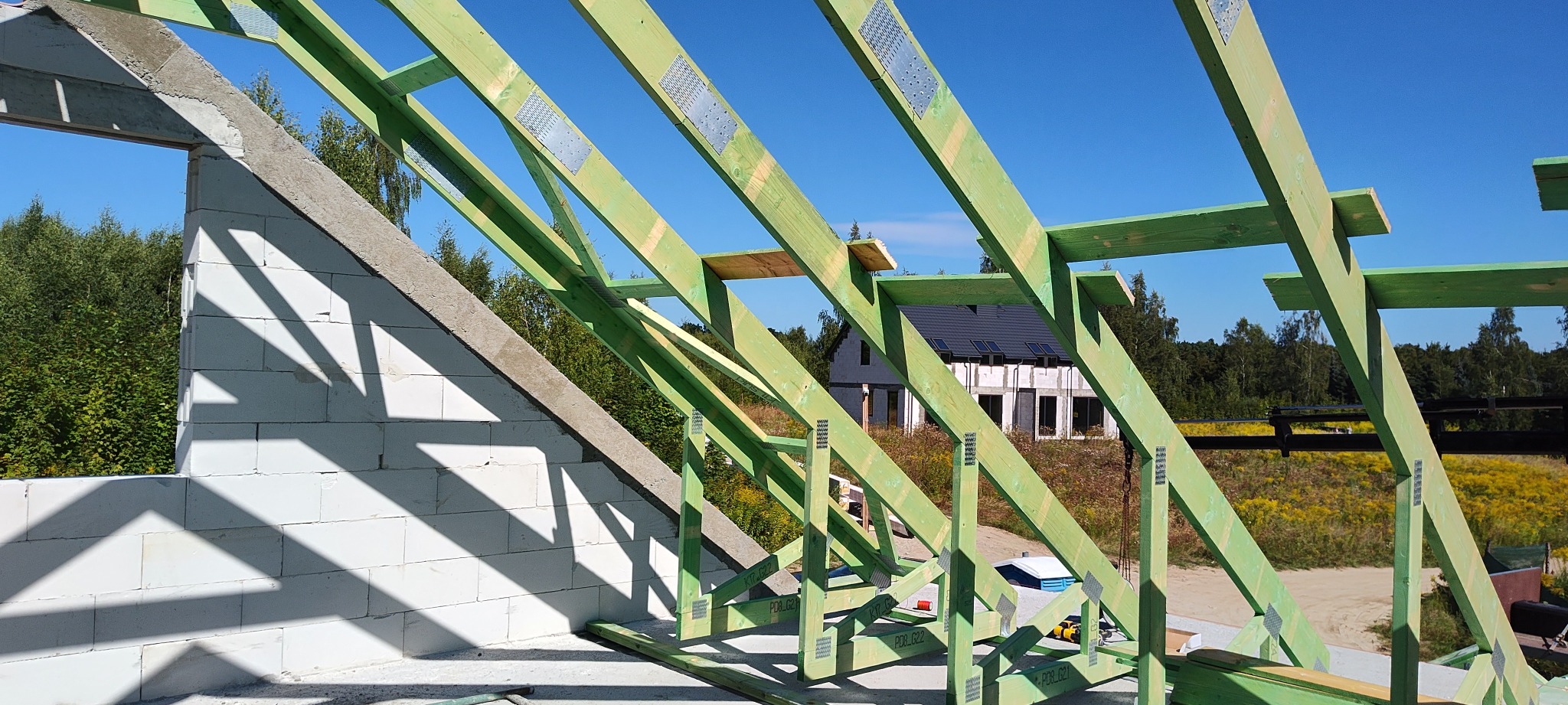 Konstrukcja dachu z zielonego drewna z metalowymi łącznikami, w tle ściana z betonu komórkowego i widok na inny dom w budowie, błękitne niebo.