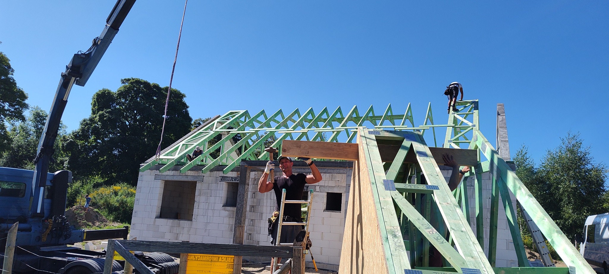 Montaż drewnianej konstrukcji dachu na budynku z białych bloczków, z użyciem dźwigu i drabiny, przy słonecznej pogodzie.