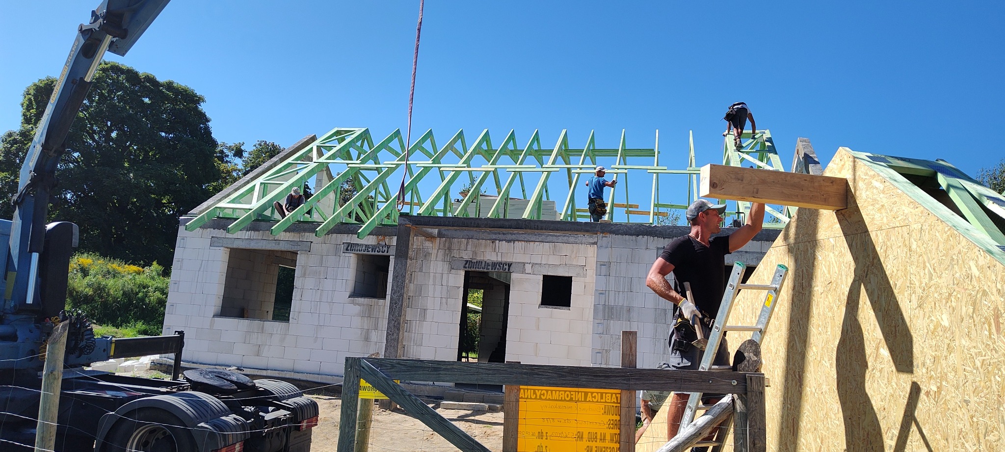 Budowa domu: robotnicy montują drewnianą konstrukcję dachu na budynku z białego bloczka, widoczny dźwig i płyta OSB na ścianie szczytowej.