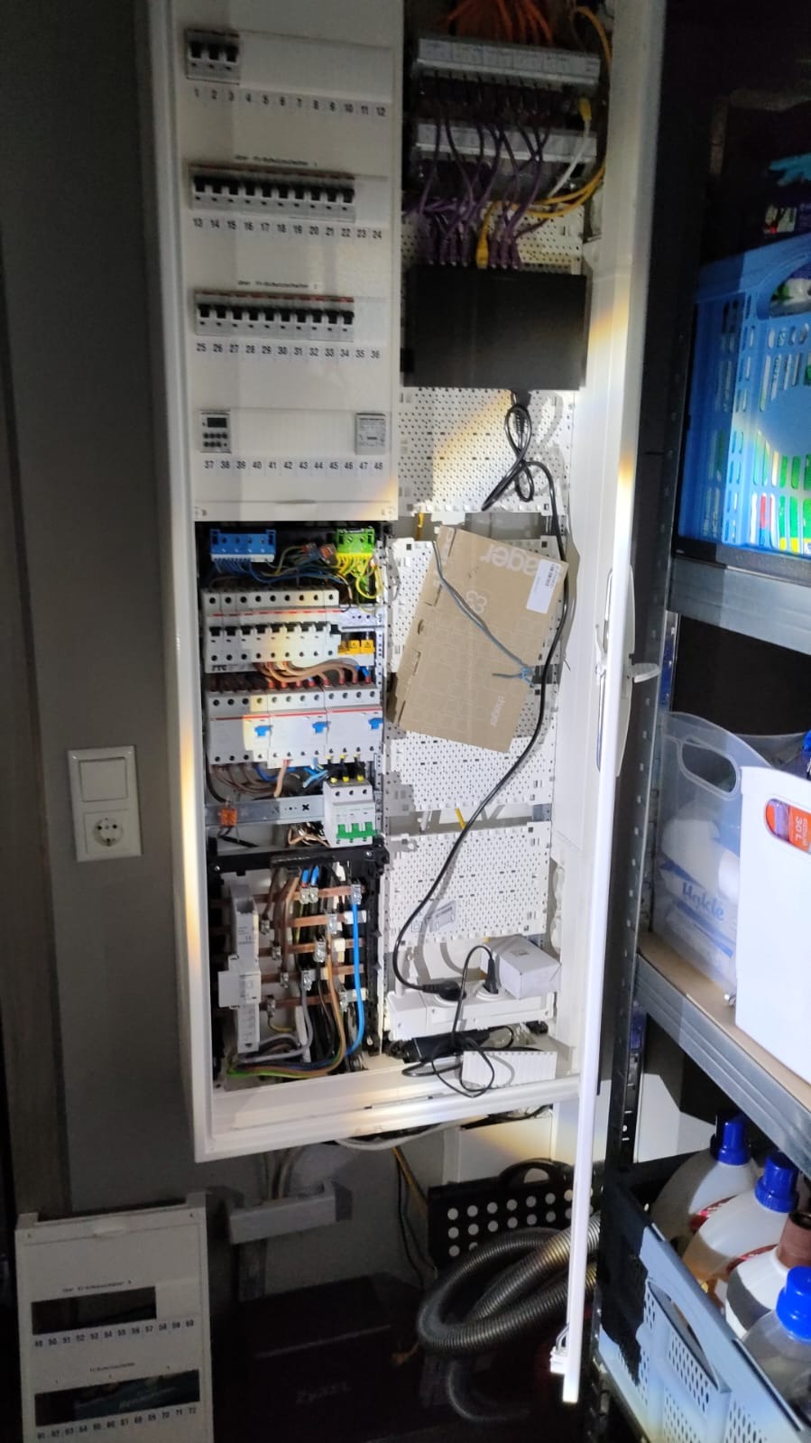 Otwarta szafa rozdzielcza z widocznymi zabezpieczeniami, przewodami i elementami sterującymi. W tle widoczne półki regału z różnymi przedmiotami. Na drzwiach szafy widoczne numery identyfikacyjne.