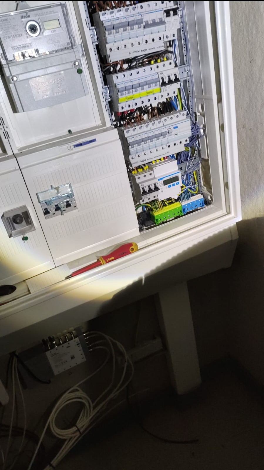 Otwarta skrzynka elektryczna z bezpiecznikami, licznikiem i okablowaniem, częściowo zakryta, z czerwono-żółtym śrubokrętem leżącym na półce; widoczny także rozdzielacz antenowy i plątanina kabli...