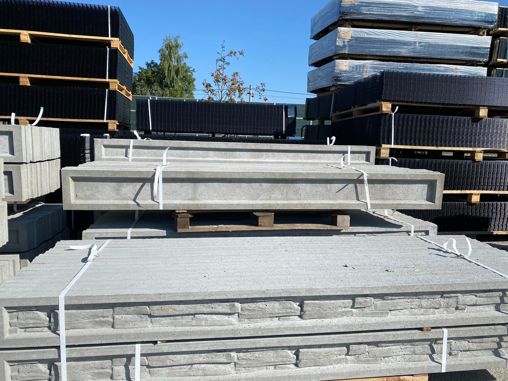 Stosy prefabrykowanych elementów betonowych z fakturą kamienia, oraz czarnych paneli ogrodzeniowych i siatki na paletach, przygotowane do transportu.