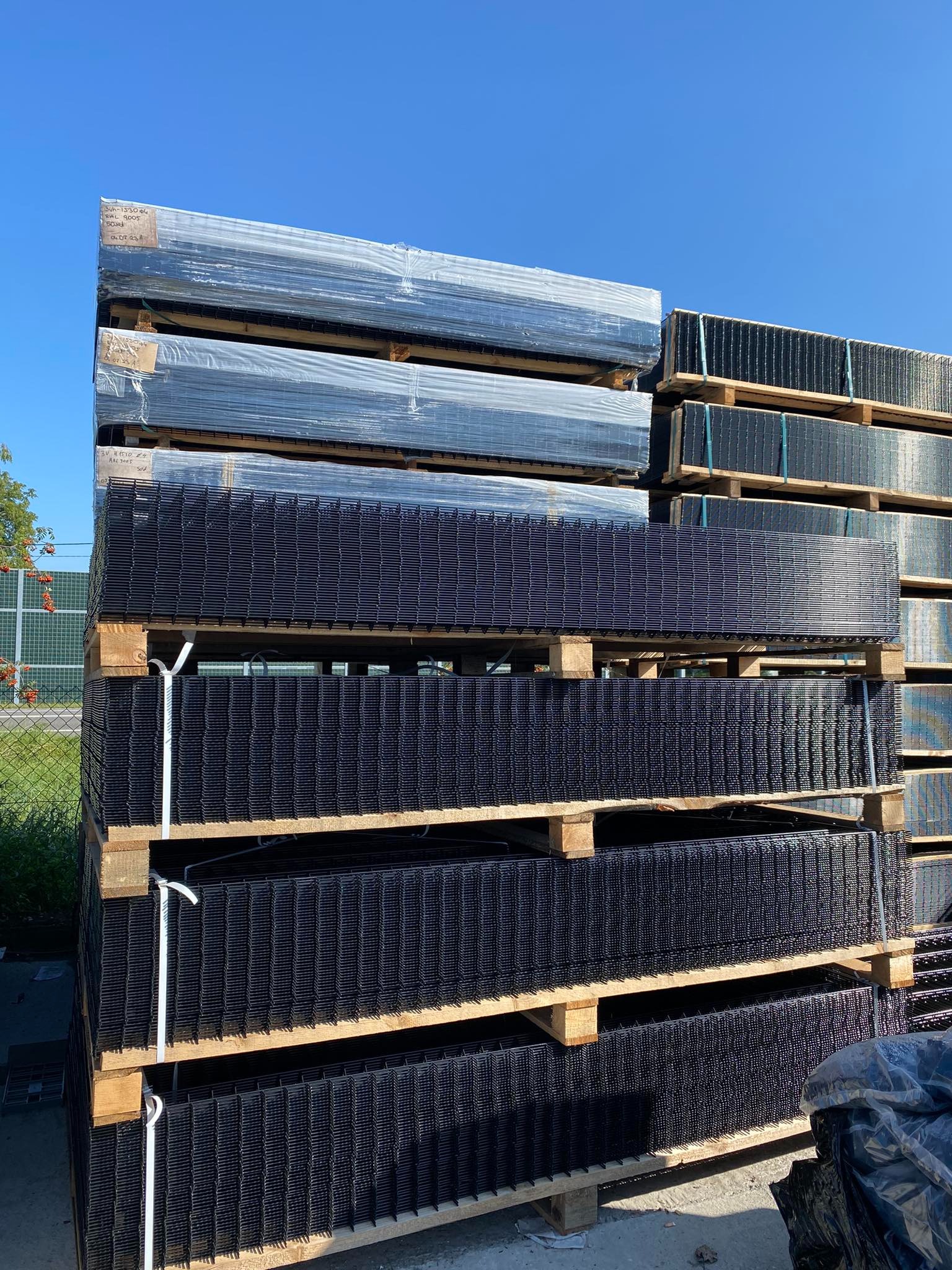 Stos palet z panelami ogrodzeniowymi, część zafoliowana, widoczne etykiety z kodami RAL 9005 i RAL 3005, błękitne niebo w tle.