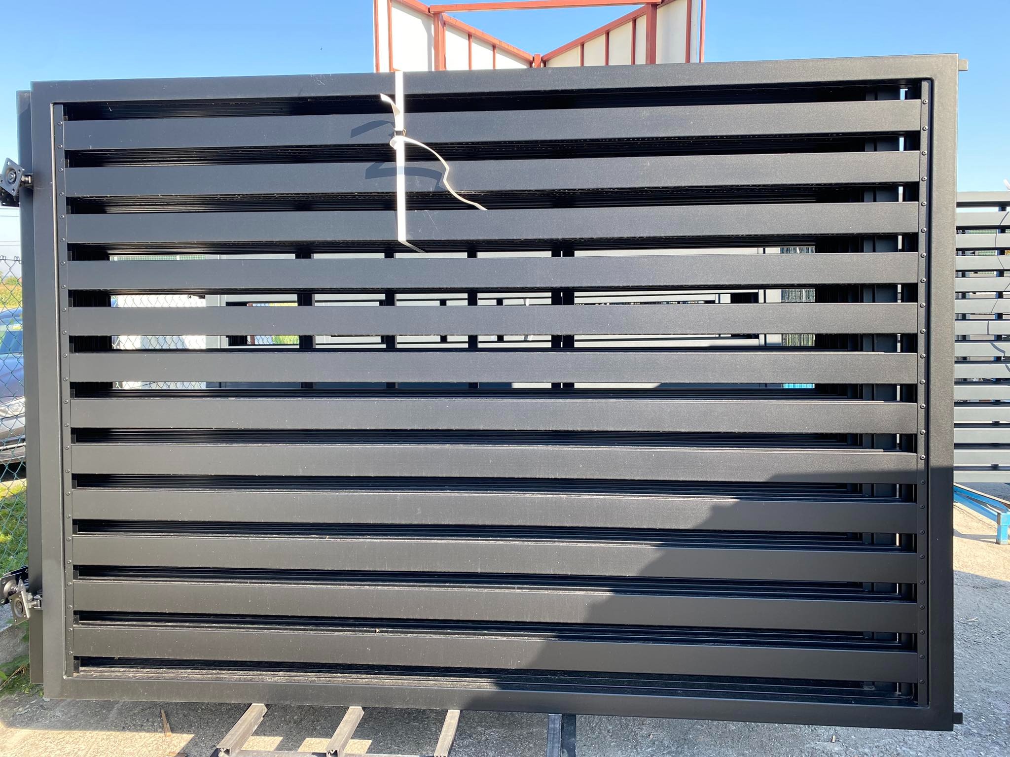 Część nowego, czarnego ogrodzenia z poziomych, metalowych profili, zabezpieczona białą linką, oczekuje na montaż.