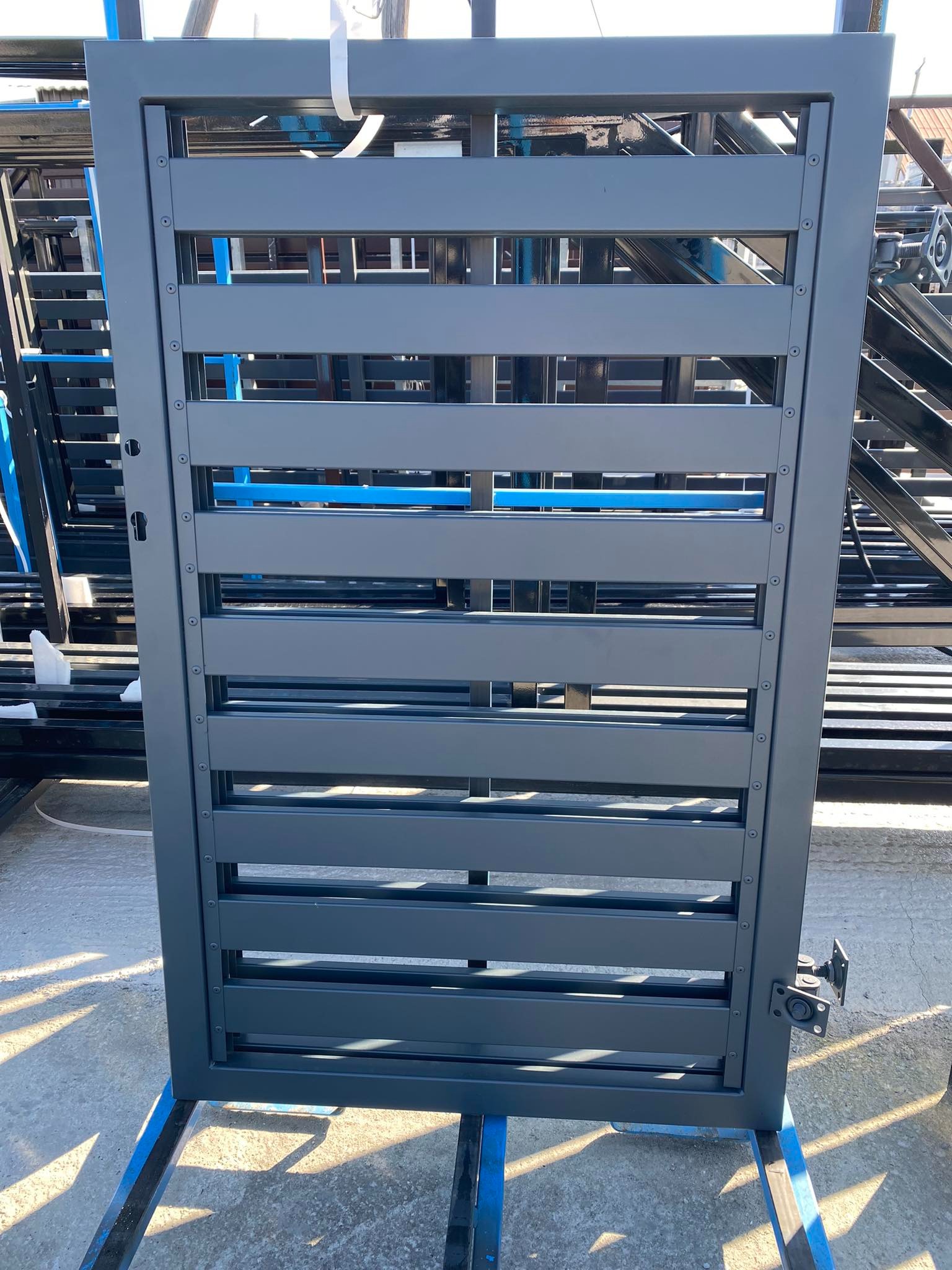 Szara, metalowa furtka z poziomymi panelami, widoczne zawiasy i zamek, ustawiona na niebieskim stelażu.