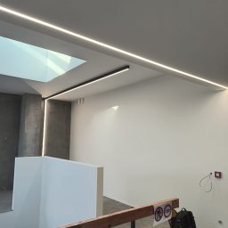 Electro-Instal - Nowoczesne oświetlenie LED wzdłuż sufitu i ściany, z widocznym świetlikiem. Minimalistyczny design, jasne światło, surowe wykończenie wnętrza, torba narzędziowa w rogu.