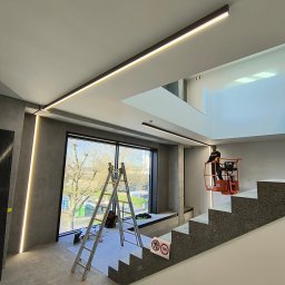 Electro-Instal - Montaż podłużnego oświetlenia LED w nowoczesnym wnętrzu. Pracownik na podnośniku, drabina, laser krzyżowy. Duże okno z widokiem na drzewa.