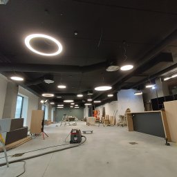 Electro-Instal - Sufit z zamontowanym oświetleniem LED w kształcie okręgów w przestrzeni komercyjnej w trakcie wykańczania. Widoczne narzędzia i elementy wyposażenia.