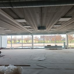 Electro-Instal - Wnętrze w trakcie prac instalacyjnych: surowy betonowy strop z zamontowanym oświetleniem, duże okna z widokiem na zewnątrz, materiały budowlane na podłodze.
