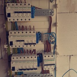 Electro-Instal - Rozdzielnica elektryczna z zainstalowanymi bezpiecznikami i przewodami wewnątrz, zamontowana na ścianie z surowej cegły. Widoczne elementy marki Schneider Electric i Smitec.