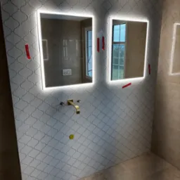 Łazienka w trakcie wykańczania: ściana pokryta białą mozaiką o marokańskim wzorze, dwa podświetlane lustra z czerwonymi klinami dystansowymi, złota bateria umywalkowa bez podłączonej umywalki...