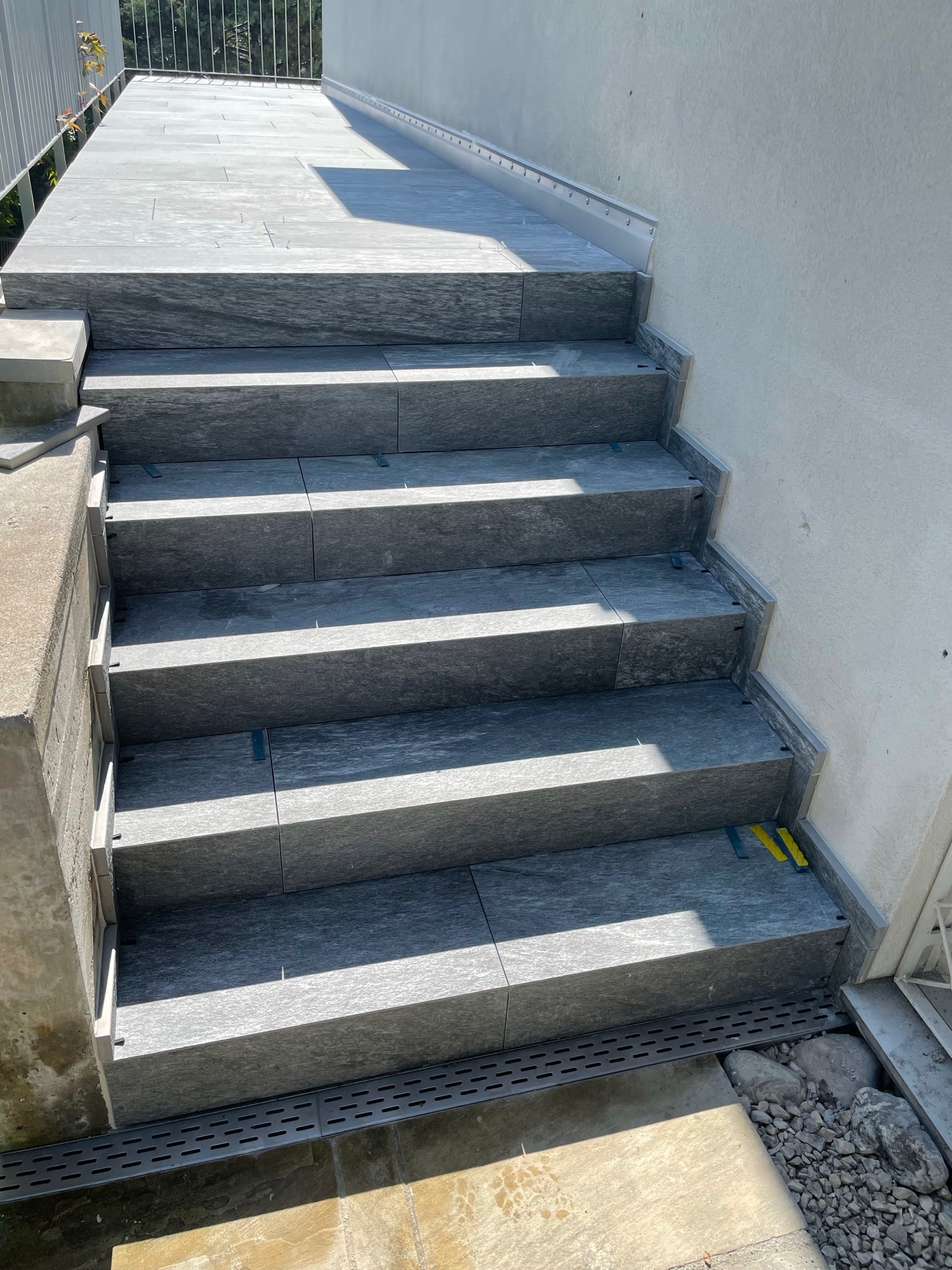 Schody zewnętrzne wyłożone szarymi płytkami imitującymi kamień, z widocznymi elementami mocującymi i niewielkimi żółtymi podkładkami dystansowymi, prowadzące do jasnej ściany.