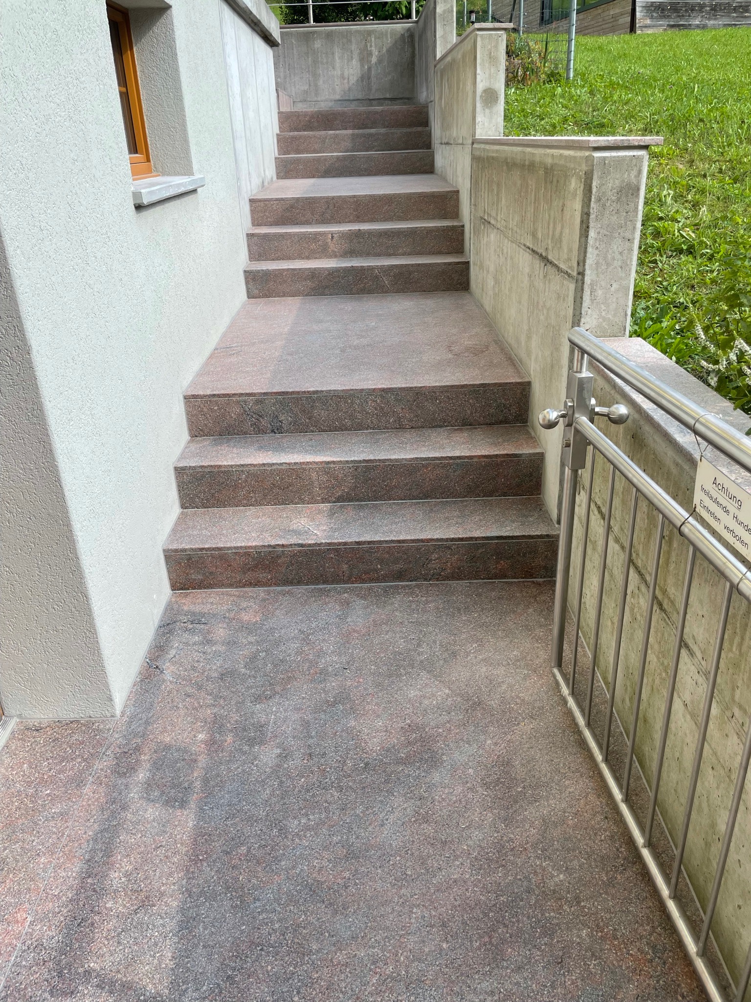 Zewnętrzne schody z czerwonego granitu prowadzące w górę, z betonowymi ścianami po bokach i metalową barierką po prawej stronie z tabliczką informacyjną.