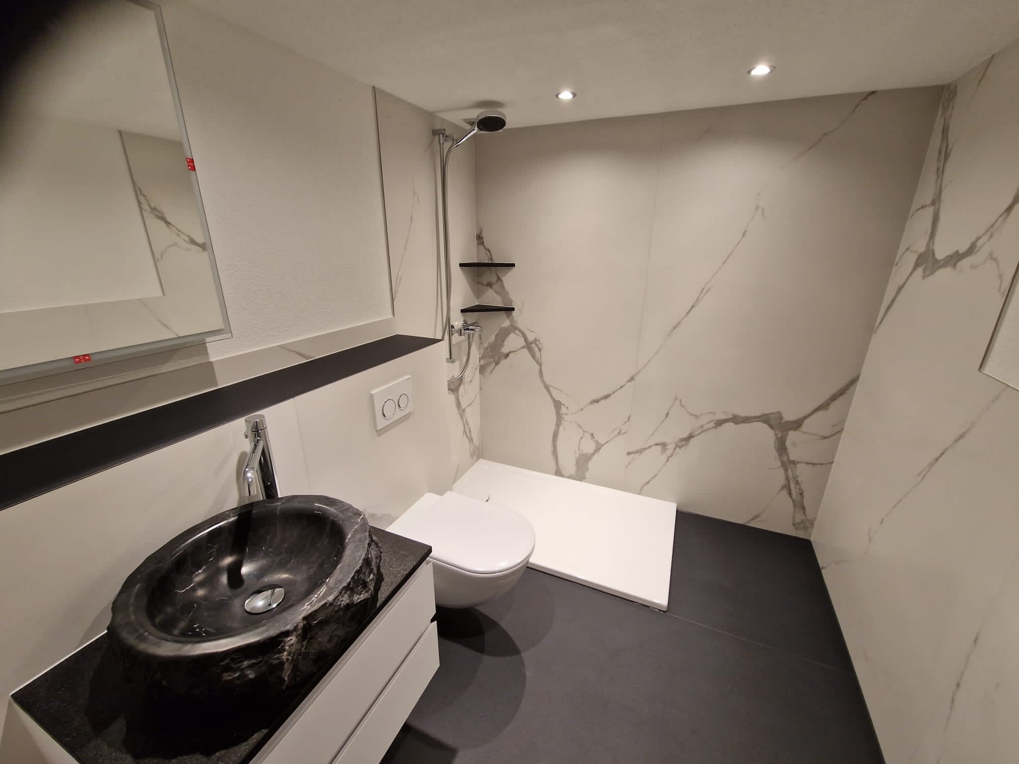 Nowoczesna łazienka z czarną kamienną umywalką, białymi płytkami imitującymi marmur, bezprogowym prysznicem i czarną podłogą.