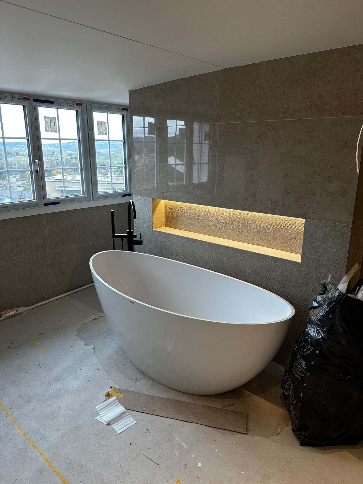 Wolnostojąca wanna z czarną armaturą na tle ściany z płytek i podświetlanej niszy w łazience w trakcie wykańczania w Rabce-Zdroju, widoczne okna z widokiem na miasto.