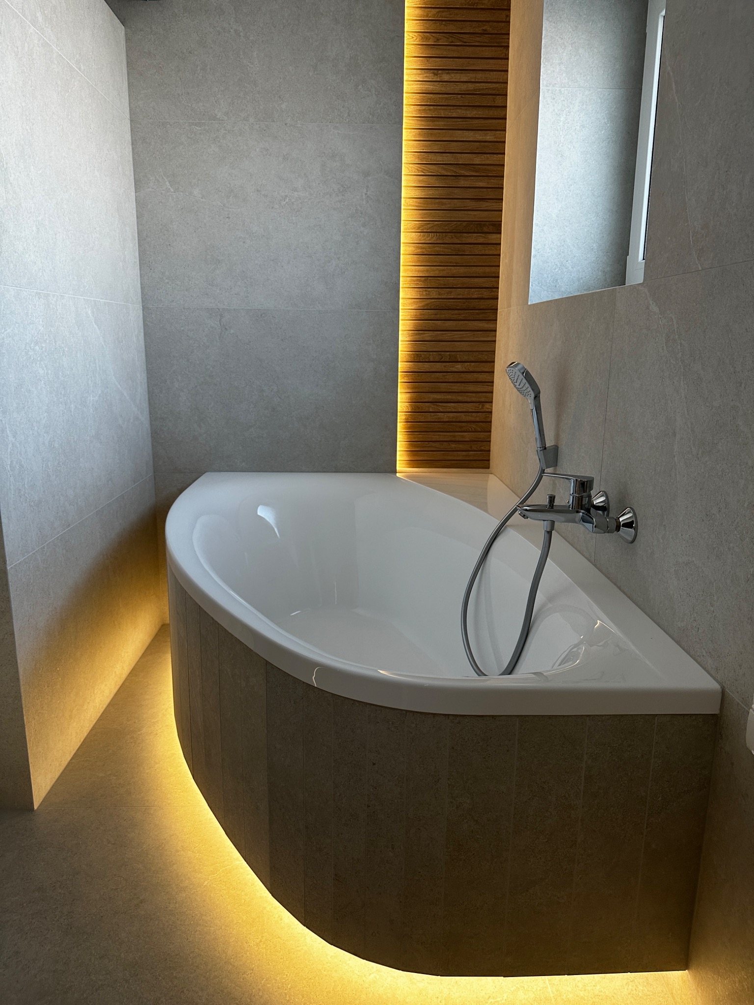 Narożna wanna obudowana płytkami w odcieniach beżu, podświetlona od dołu ciepłym światłem LED, z chromowaną baterią i słuchawką prysznicową na tle szarych płytek ściennych i drewnianej dekoracji...